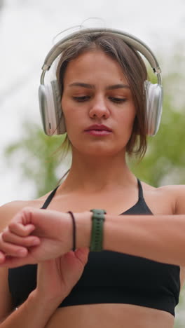 close view of focused young lady standing outdoors, wearing headphones and checking time on her green wristwatch while black bangle on hand under bright natural setting