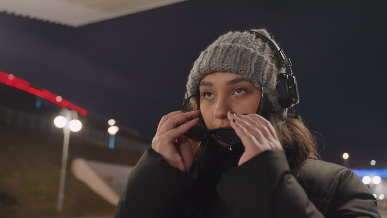 夜の水辺のダンサーがヘッドホンとサングラスを調整する。ビーニー帽とパフコートを着た若い女性が、橋の照明の下、遊歩道に立つ。襟を立て、サングラスをかけると、自信に満ちたストリートの雰囲気に変わる。