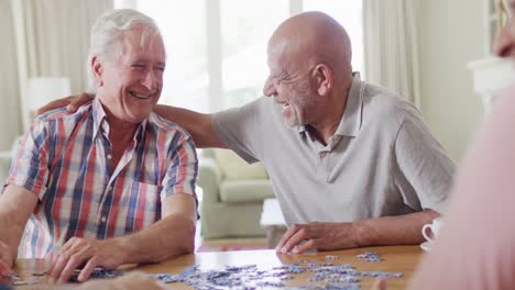 Two-diverse-senior-male-friends-embracing,-doing-jigsaw-puzzle-in-living-room,-slow-motion