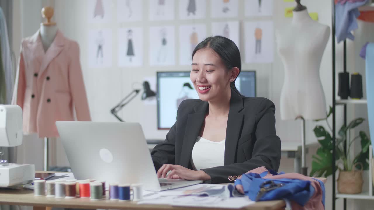 사업복을 입은 미소 짓는 아시아 여성 디자이너가 노트북에서 일하고 스튜디오에서 옷을 디자인하는 동안 커피를 마시고 있습니다.