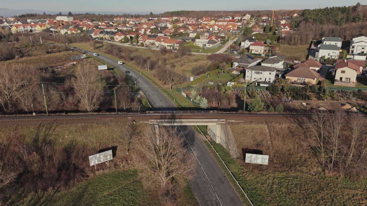 Letecký pohled na jasně žlutozelený osobní vlak přejíždějící most nad venkovskou silnicí na předměstí. Železnici obklopují domy, klikaté ulice a otevřená pole.
