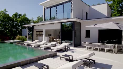 maravillosa vista de una lujosa villa negra y blanca desde su gran terraza con una piscina infinita, muchas tumbonas, un balcón y ventanas de piso a techo, clima soleado