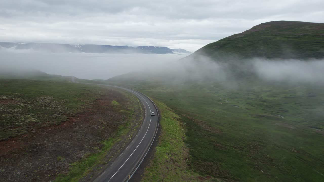 Aerial view of a car driving through foggy mountains along the Ring Road in Iceland during summer. The vehicle winds through mist-covered peaks, surrounded by dramatic landscapes and low visibility