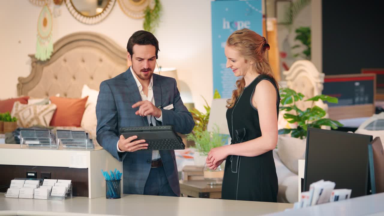 A Young Man Gestures for a Female Customer to Look at a Sales Tablet in a Furniture Store