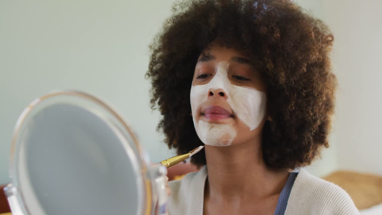 mujer biracial feliz aplicando máscara facial de belleza con pincel