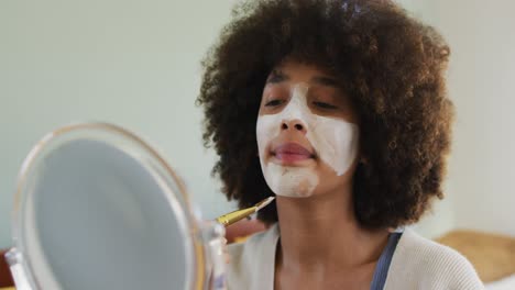 mulher biracial feliz aplicando máscara facial de beleza com escova