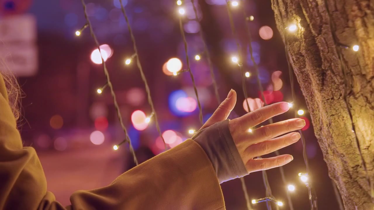 White woman hand tracing warm lights with fingerless glove and coat sleeve visible, closeup on tactile interaction, bulbs casting amber bokeh, gentle motion, intimate holiday detail, illumination