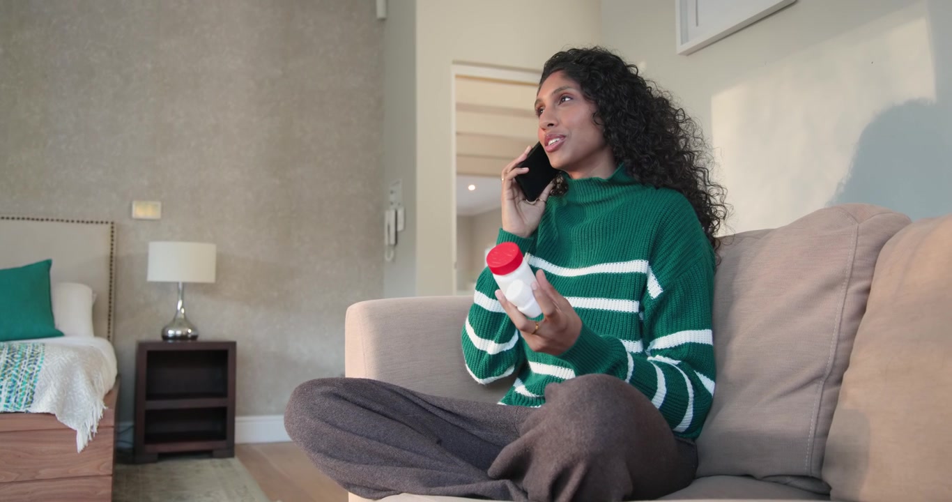 Afroamerikansk kvinde modtager opkald, holder smartphone til øret på sofaen og sammenligner pilleflasker.