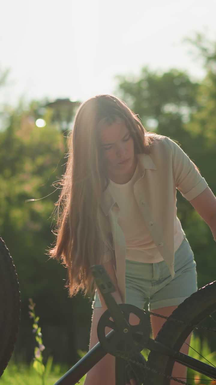 Young woman slightly bent adjusts front tire of her upside-down bike while rotating back tire, hair flowing down, sunlight reflecting off her, surrounded by lush greenery, background includes trees