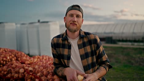 Portrait-of-a-happy-male-farmer-in-a-plaid-shirt-who-is-leaning-on-bags-of-onions-after-a-successful-harvest-season-on-the-farm