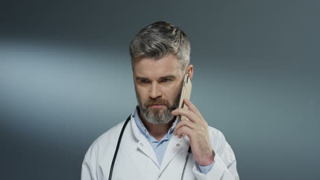 good looking grey haired male medic in white gown speaking on the mobile phone on the gray background