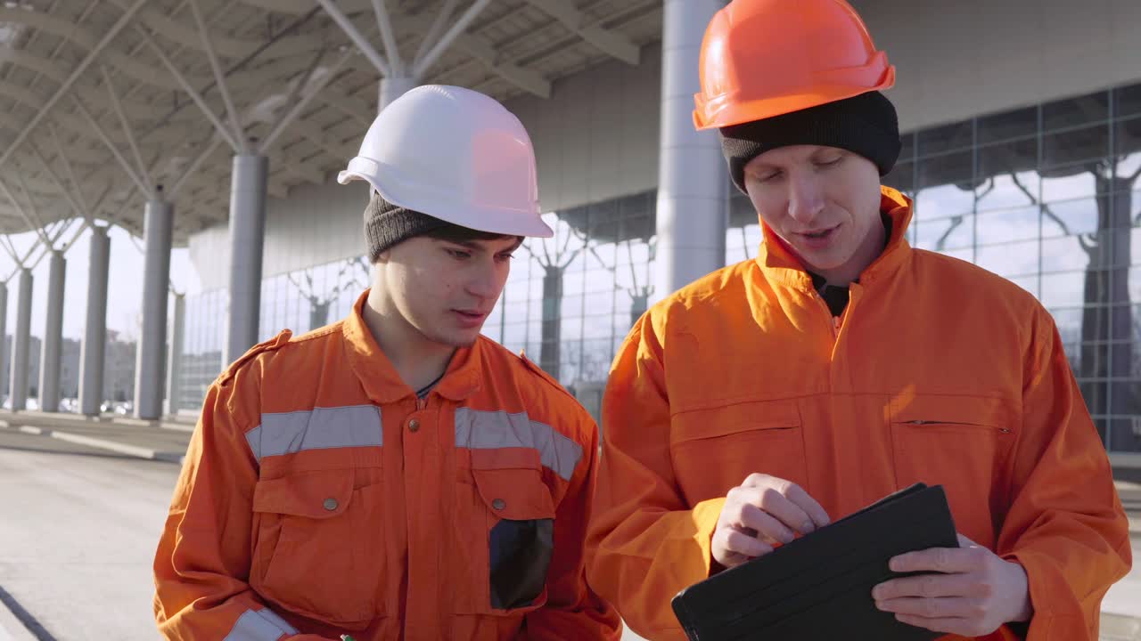 Two construction workers in orange uniform and helmets walking through the construction field and looking over plans together. Building at the background