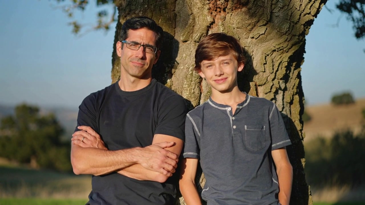 La telecamera si stringe, padre con gli occhiali e figlio appoggiati a un tronco d'albero, in posa su un pendio erboso.