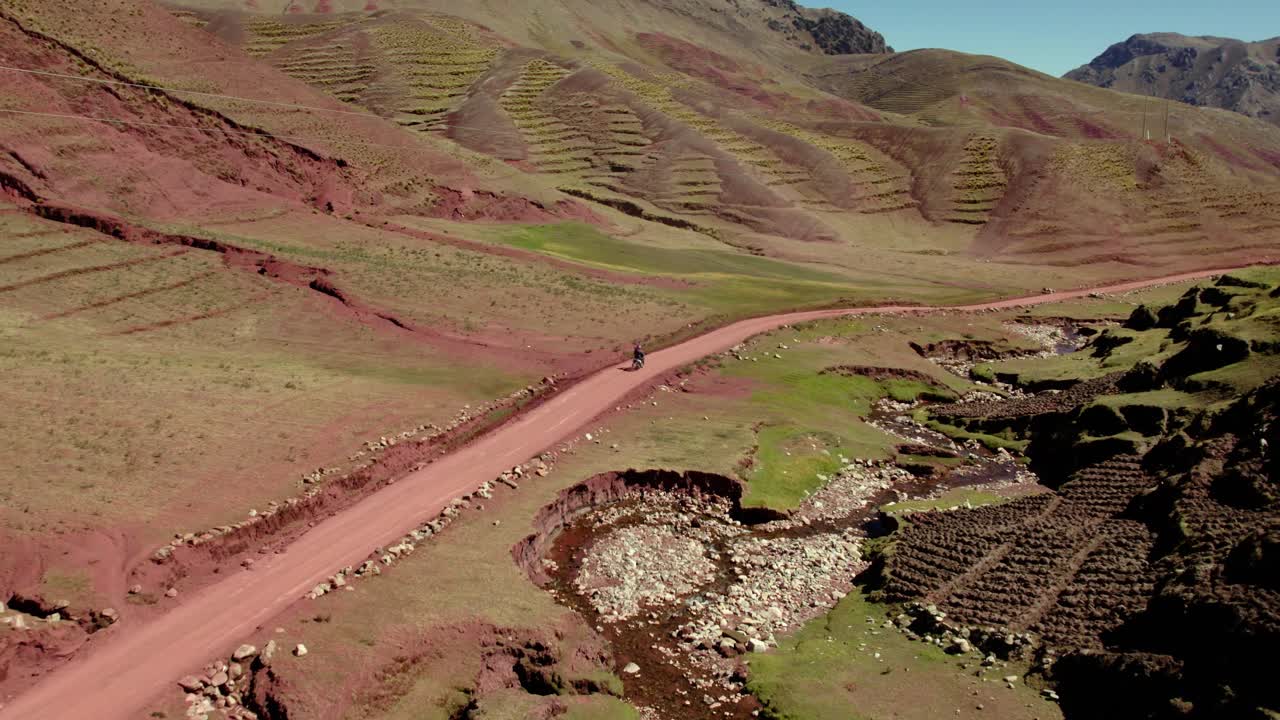 un motociclista montando en un sinuoso camino de tierra a través de tierras de cultivo en terrazas en una región montañosa. el sol brilla brillantemente, proyectando sombras en los campos verdes exuberantes y el terreno accidentado.