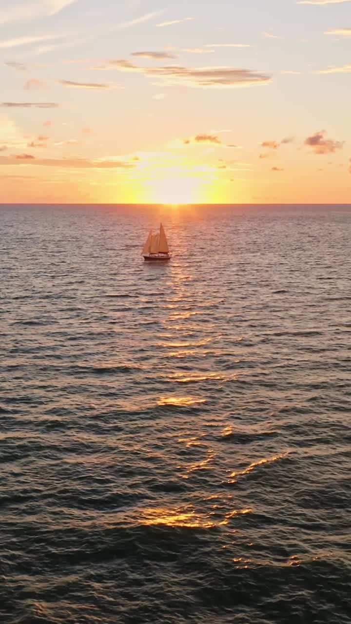 A lone sailboat moves steadily across the sea as the sun sets on the horizon, scattering golden reflections across the rippling waves in a tranquil evening scene