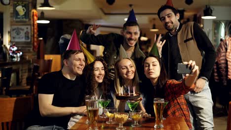 amigos felices están haciendo selfies con pastel de cumpleaños usando teléfonos inteligentes mientras celebran en el café. los jóvenes se están divirtiendo, haciendo caras tontas y riendo.