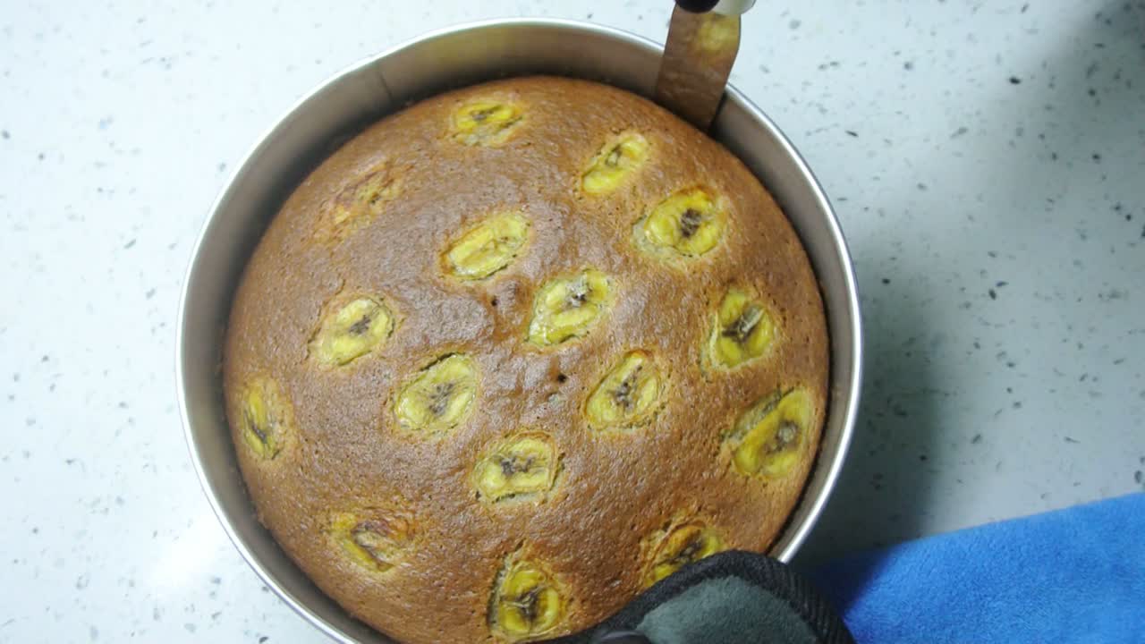 The process of removing the banana cake from a baking tray. The cake surface is carefully separated from the baking tray using a long spatula. The cake will be transferred to the cake board.