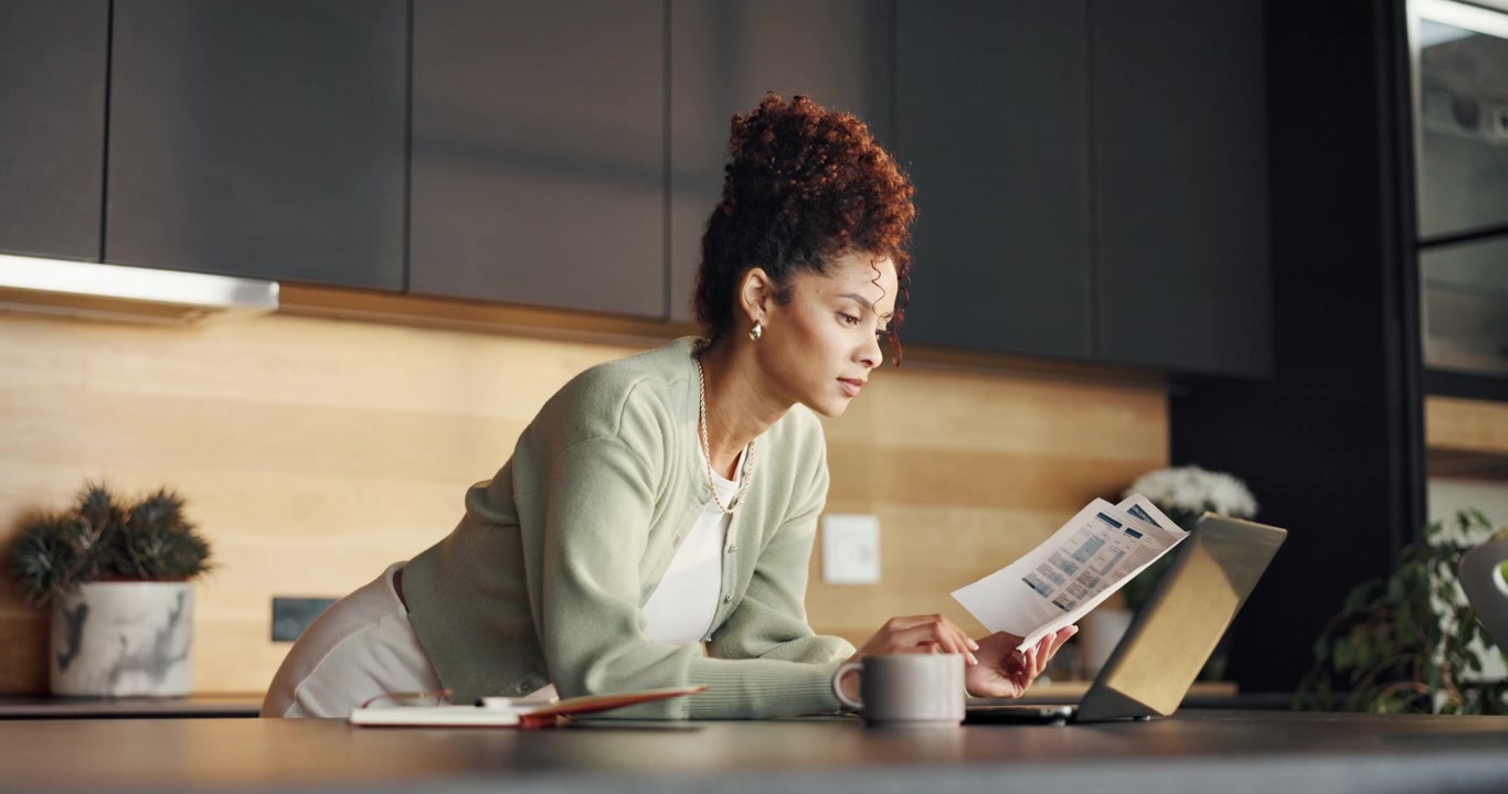 Young Woman Reviewing Documents While Working on Laptop in Modern Kitchen