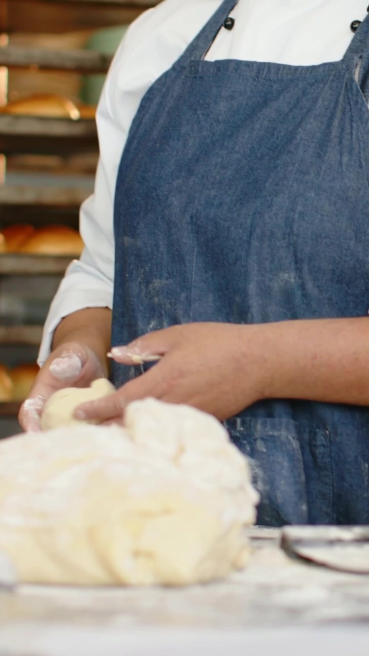 Video vertical: Panadero con chaqueta de chef y delantal vaquero alcanzando la masa y dando forma a los panecillos en la panadería junto a un estante.