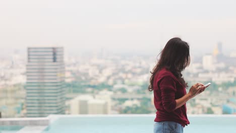 side view of concentrated woman wearing red sweater texting on a smartphone while walking alone on skyscraper near swimming pool in bright sunlight.