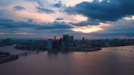 stunning drone footage captures phnom penh's skyline at dusk, with skyscrapers silhouetted against vibrant sunset hues. the calm river reflects the dramatic cloudscape above