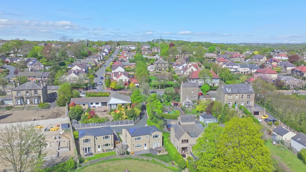 Aerial drone shot flies over treetops and rooftops, gradually revealing Shibden Park and the adjacent residential neighborhood in Halifax under a spring sky, slow motion