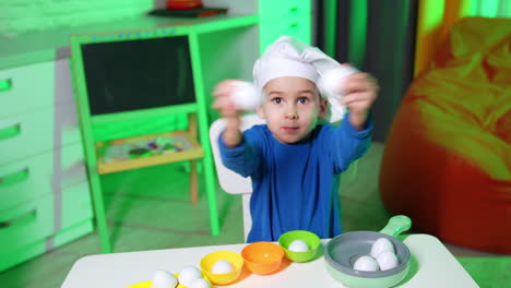 chef baby boy in white cap plays with toy eggs. happy kid puts the eggs into the colorful cups. high angle view.