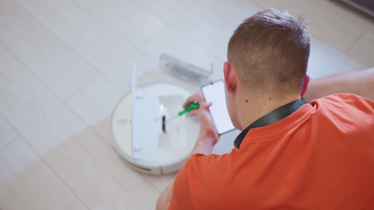 Close up rear view of engineer in orange shirt seated on floor using phone for guidance while working on opened robot vacuum, preparing tools and ensuring maintenance for better cleaning performance