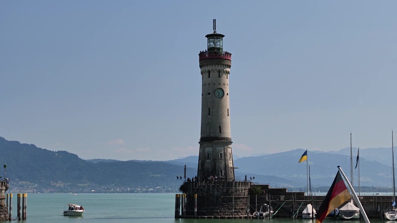 Piccola barca a motore che naviga oltre il Nuovo Faro di Lindau all'ingresso del porto sul Lago di Costanza, con montagne sullo sfondo.