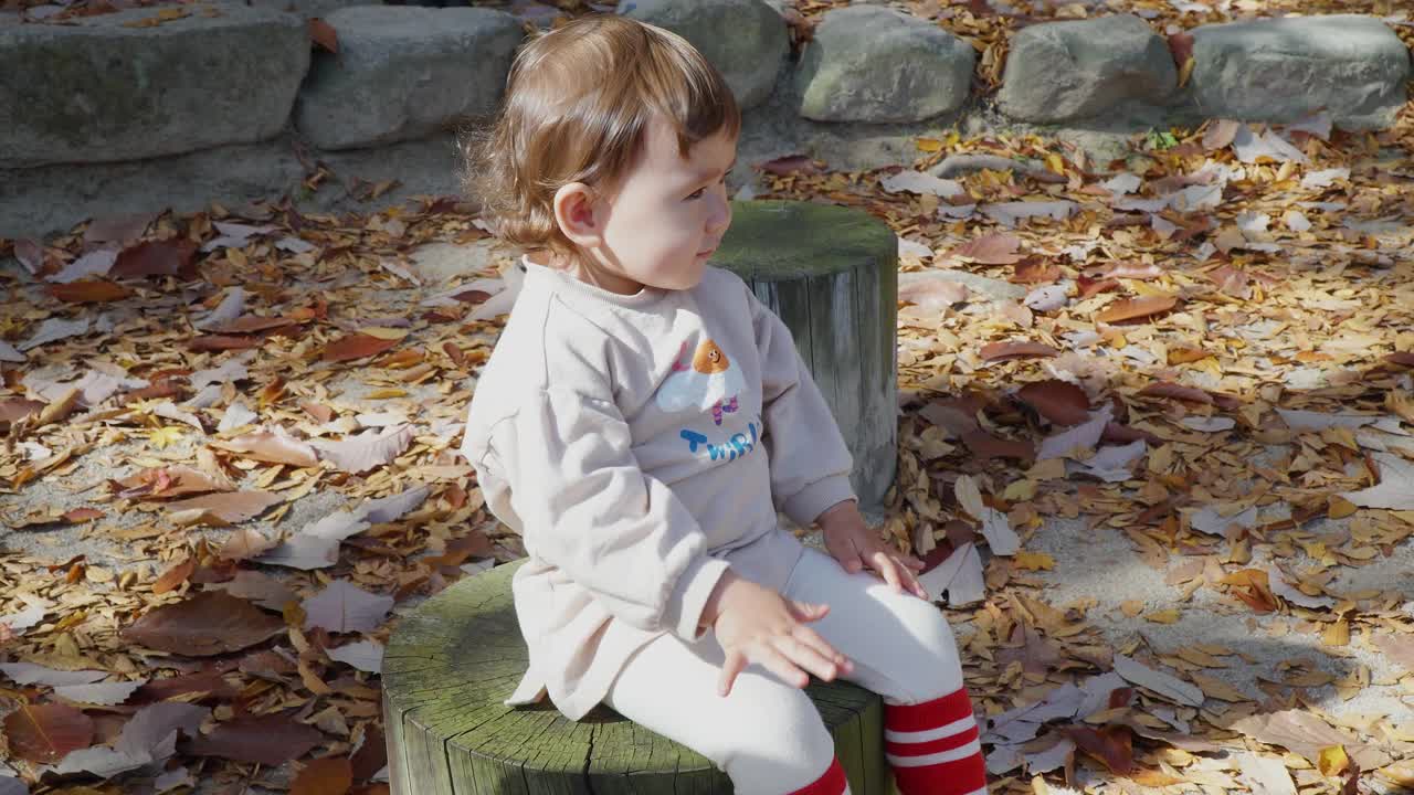 Baby toddler girl sitting on wooden stump in a park and clapping on her knees with excitement