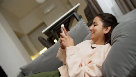 Happy-brunette-Indian-girl-in-light-pink-on-a-gray-sofa-during-her-relaxation-and-typing-on-social-networks-using-a-white-smartphone-at-home-in-a-modern-apartment