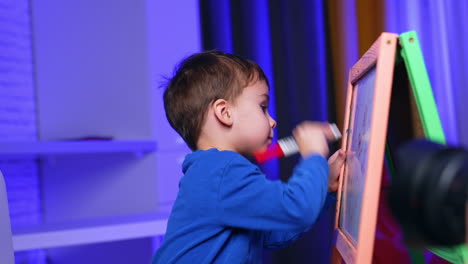 adorable dark-haired baby in blue sweater taps the blackboard with red marker. active kid claps his hand by the whiteboard and draws lines. side view.