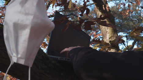 manos escribiendo laptop en el bosque durante la pandemia de covid, máscara facial colgando en el árbol, tiro cerrado