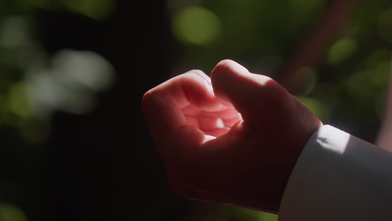 Close view of man fingers joined together against blurred green forest background, sunlight highlighting skin texture and creating artistic atmosphere of human detail in outdoor natural environment