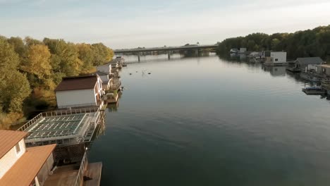 Malerische-Hausboote-In-Allen-Farben-Des-Regenbogens-Liegen-An-Einem-Flussufer,-In-Der-Ferne-Ragt-Eine-Brücke-Hervor