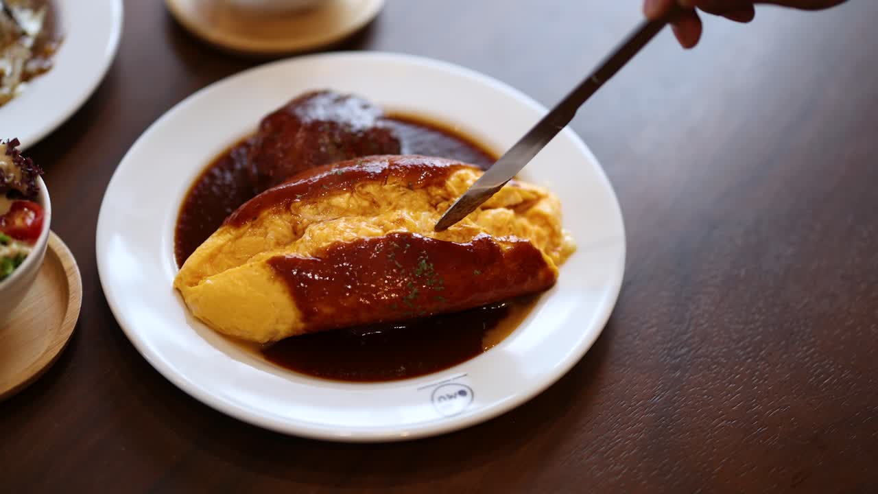 Hand slices omurice, revealing soft omelette, demi-glace sauce, and hamburg steak in natural light