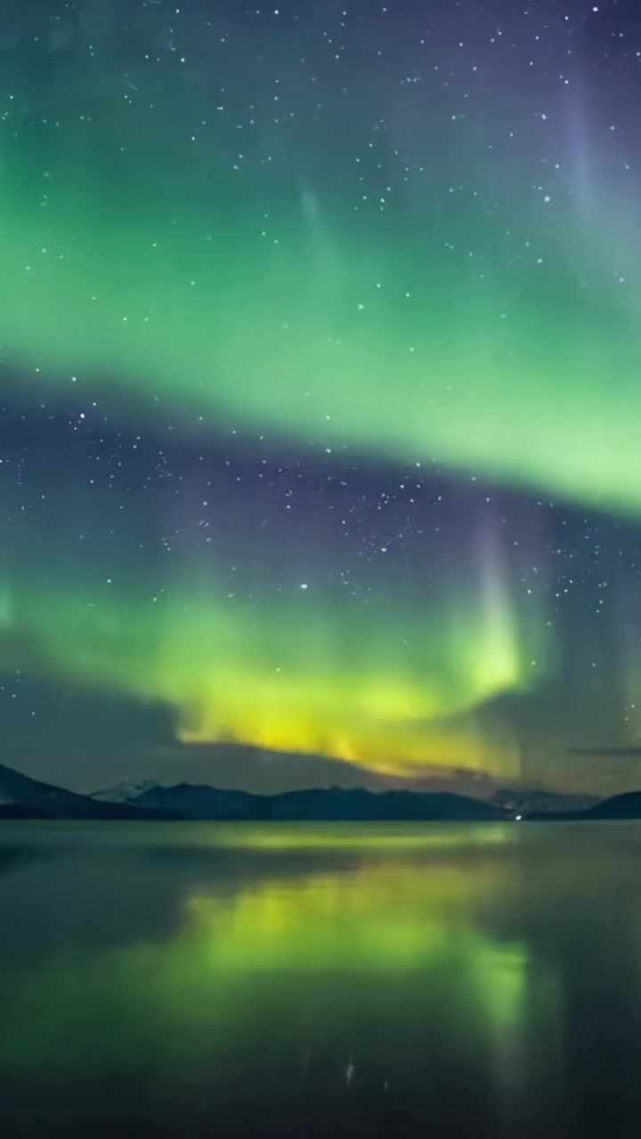 Mozzafiato Aurora Boreale che danza nel cielo notturno riflessa in un lago sereno, illuminando le stelle e le montagne in uno spettacolare display celeste.
