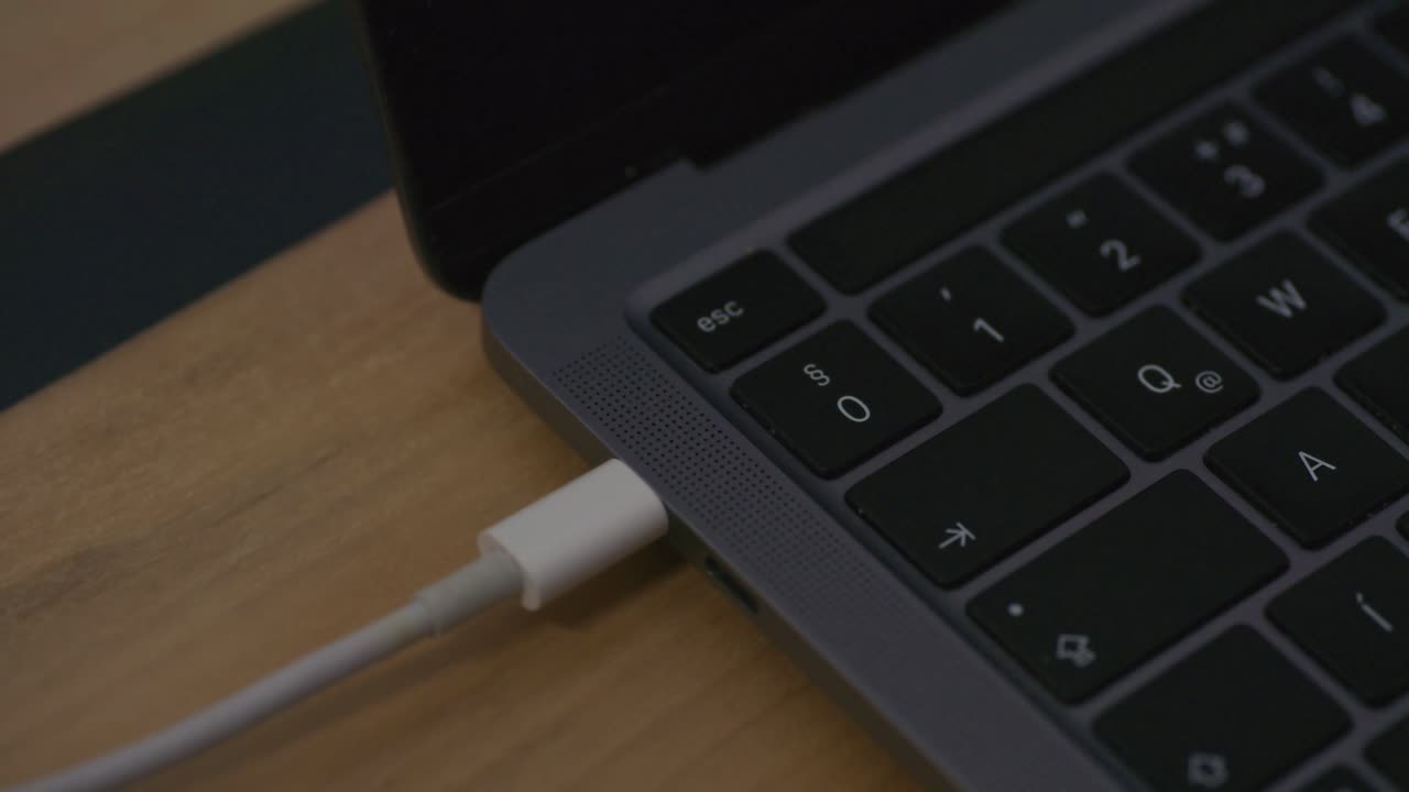 A close-up view of a person connecting a white USB-C cable to a modern laptop. The video highlights the sleek keyboard design and the precision of the charging or data transfer process