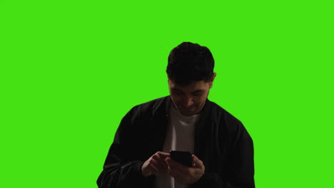 Young-Man-Looking-At-Text-Message-Or-Browsing-Online-On-Mobile-Phone-Standing-Against-Green-Screen-Background-With-Low-Key-Lighting