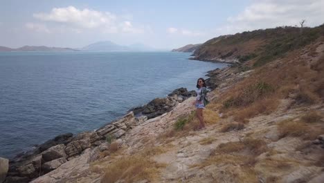 aerial: a low altitude dolly in shot of a girl standing at a high hill looking at a rocky shore and the ocean, while correcting her hair