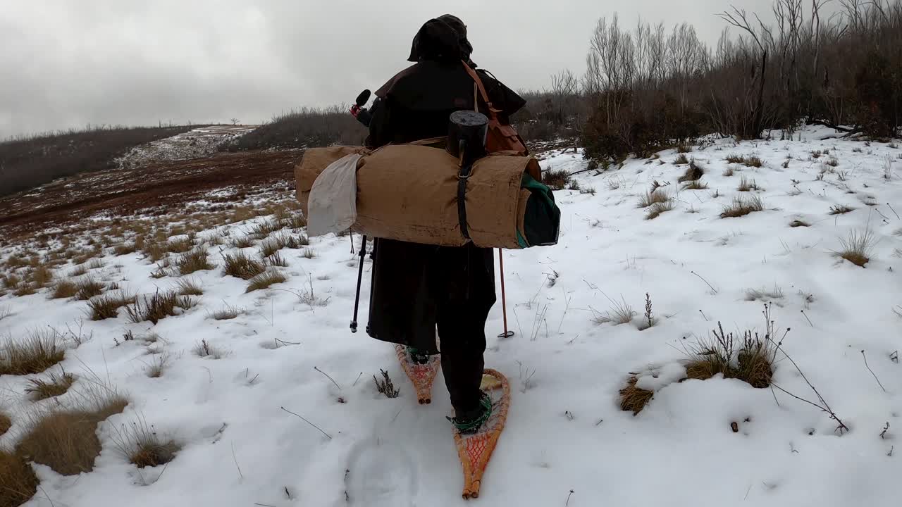 A tracking shot from behind of a man dressed as an 1800's explorer hiking through the snow in traditional clothing and camping gear