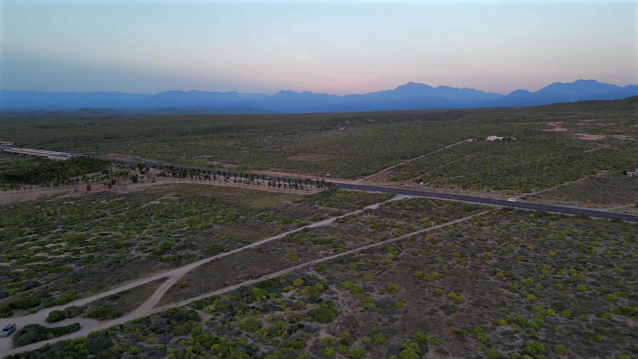 Drone slowly pans left over a lush green desert near Secret Rocks Beach below Todos Santos, watching cars pass on a distant highway beneath a mountain horizon glowing in a flat purple hue.