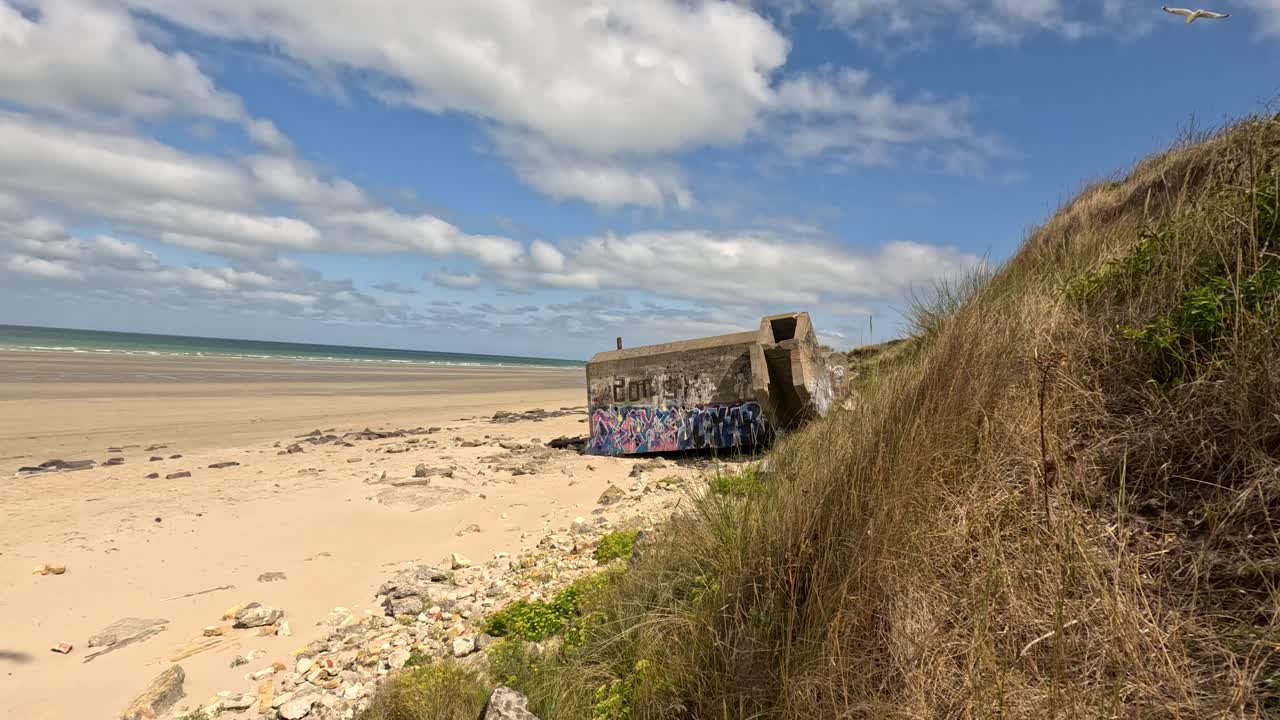 Camera smoothly pans across sandy beach and grassy dunes, revealing a graffiti-covered World War-era bunker under bright daylight at Dunkirk, France