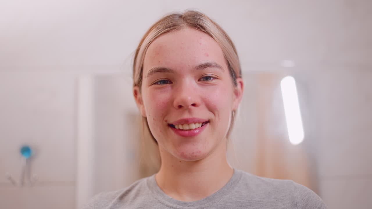 Elegant young woman standing in bathroom looking directly at camera with natural expression before smiling warmly, highlighting confidence, selfcare and positive energy