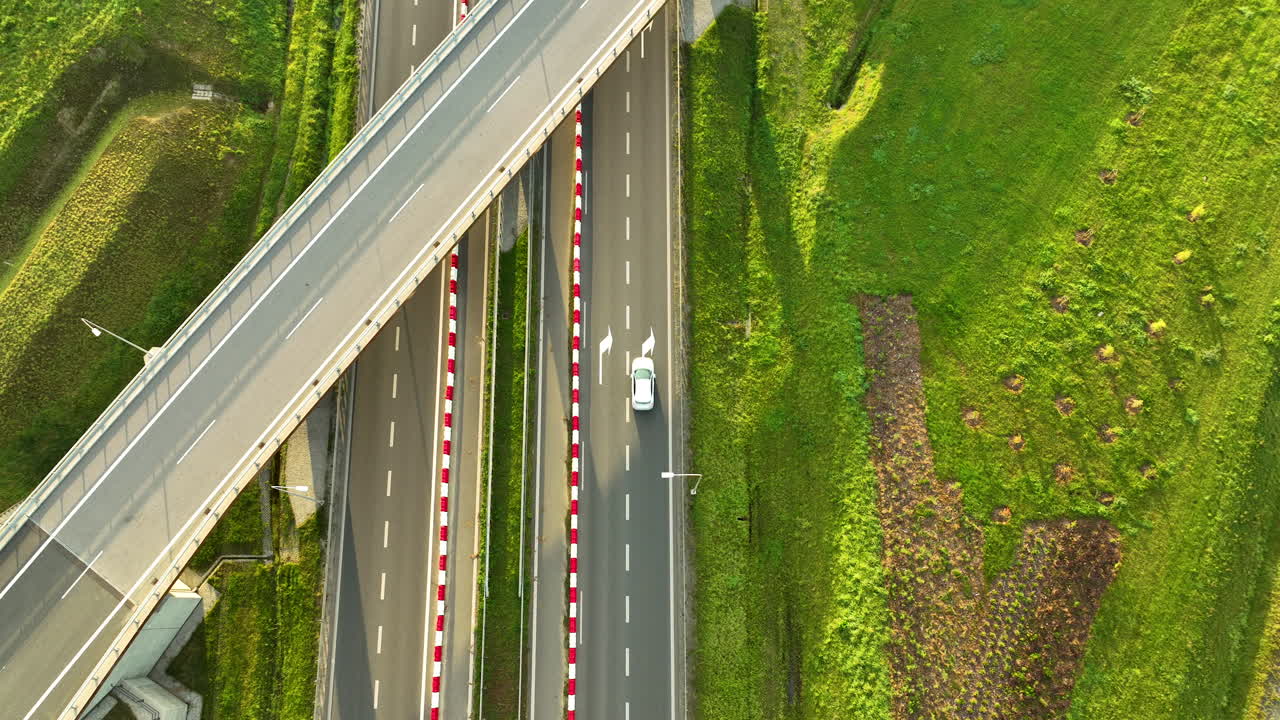 Top-down aerial shot of a white car driving under a highway overpass on the S6 expressway, highlighting the stark geometric contrast and bright green slopes