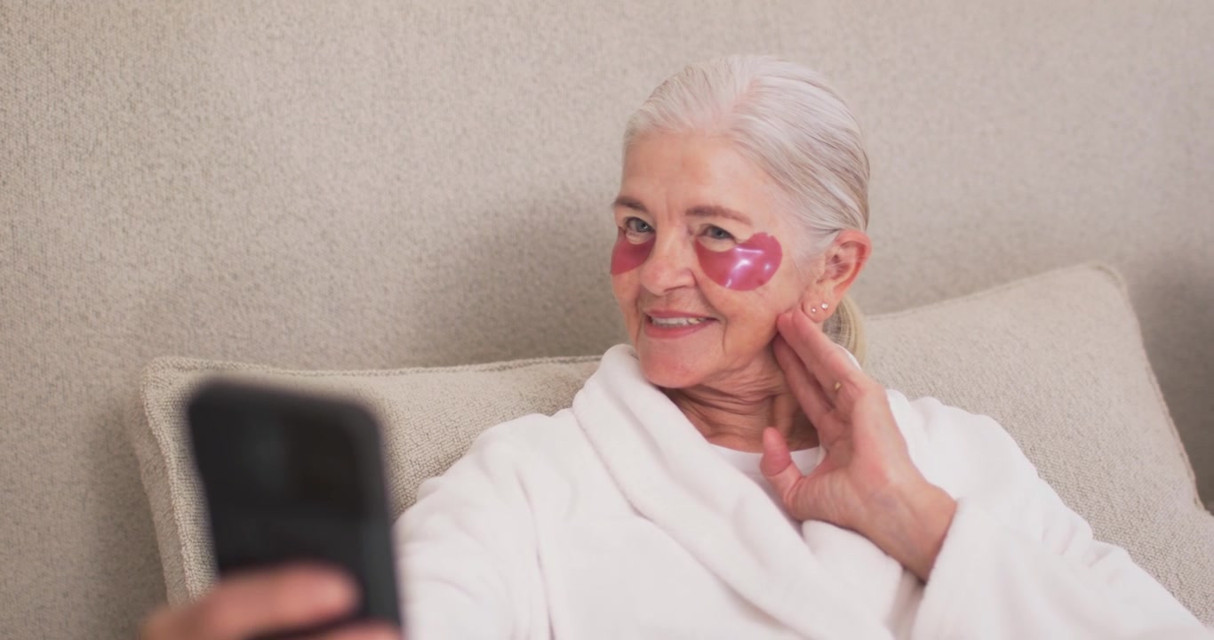 Senior woman sitting on sofa in robe seeing phone left foreground, inspecting pink heart patches