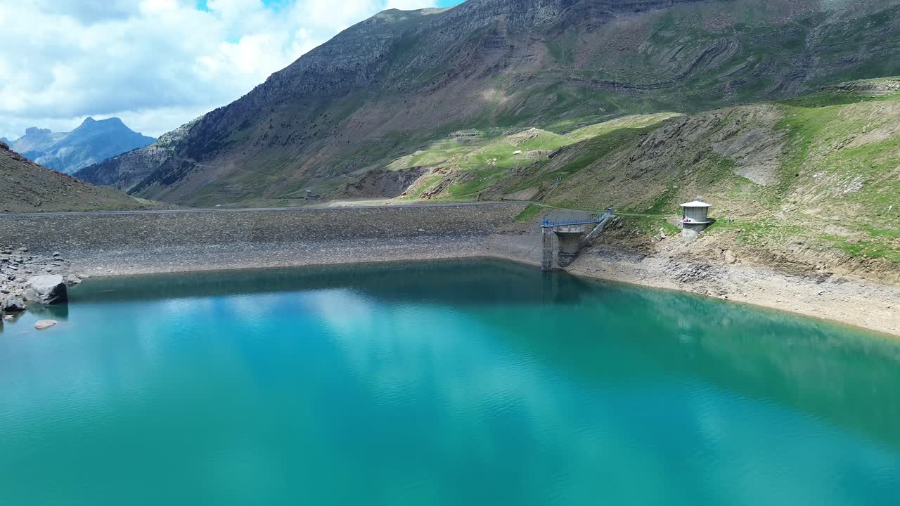 Aerial footage captures Valle de Ip's tranquil dam with azure waters nestled in the rugged mountains near Canfranc, Aragón. Bright sunlight highlights the serene landscape