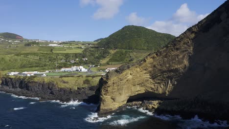 collapsed-volcano-caldera-revealing-a-village,-skate-park,-sports-field,-with-mountains,-vegetation-and-lava-formations-next-to-the-sea-drone-footage,-Velas,-Sao-Jorge-island,-Azores,-Portugal