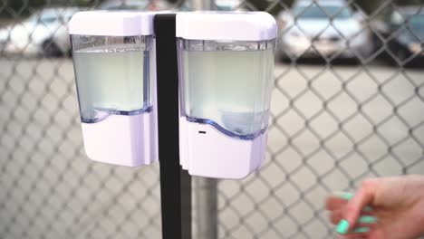 a woman using an automatic gel hand sanitizer dispenser station outside at a park in front of a chainlink fence during the covid19 coronavirus health pandemic