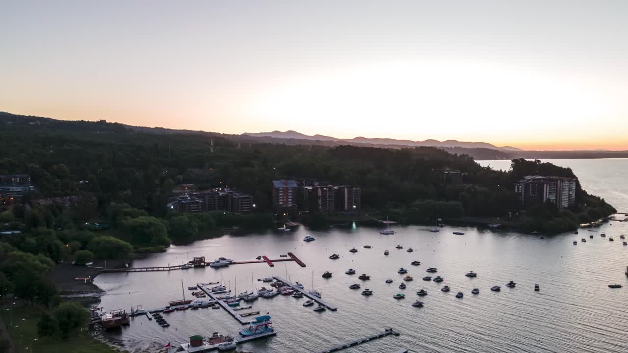 Orbit hyperlapse of boats sailing in lake near docks on green hillside, Villarrica volcano in background at sunset, La Poza, Pucon, Chile